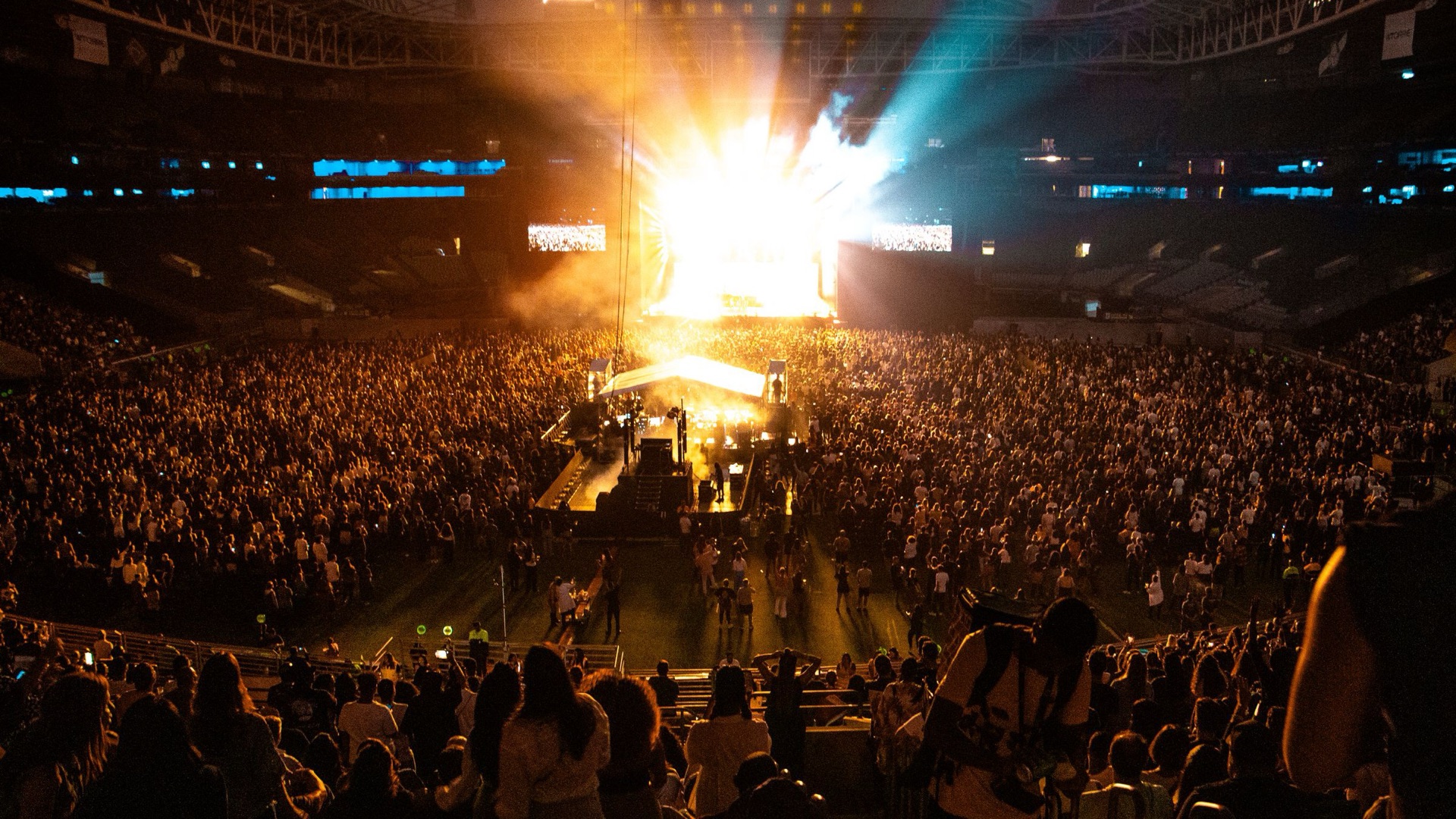 Pubblico di un concerto in uno stadio, di sera, con il palco illuminato sullo sfondo